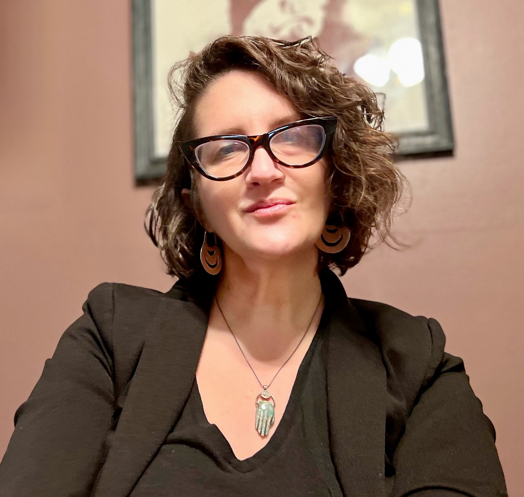 Image of Dr. Alaina Brenick in a black top and black blazer with tortoise shell glasses in a cat eye shape. She is wearing a light blue batique Hamsa necklace. She has short brown wavy hair, slightly shaved on the right side. 