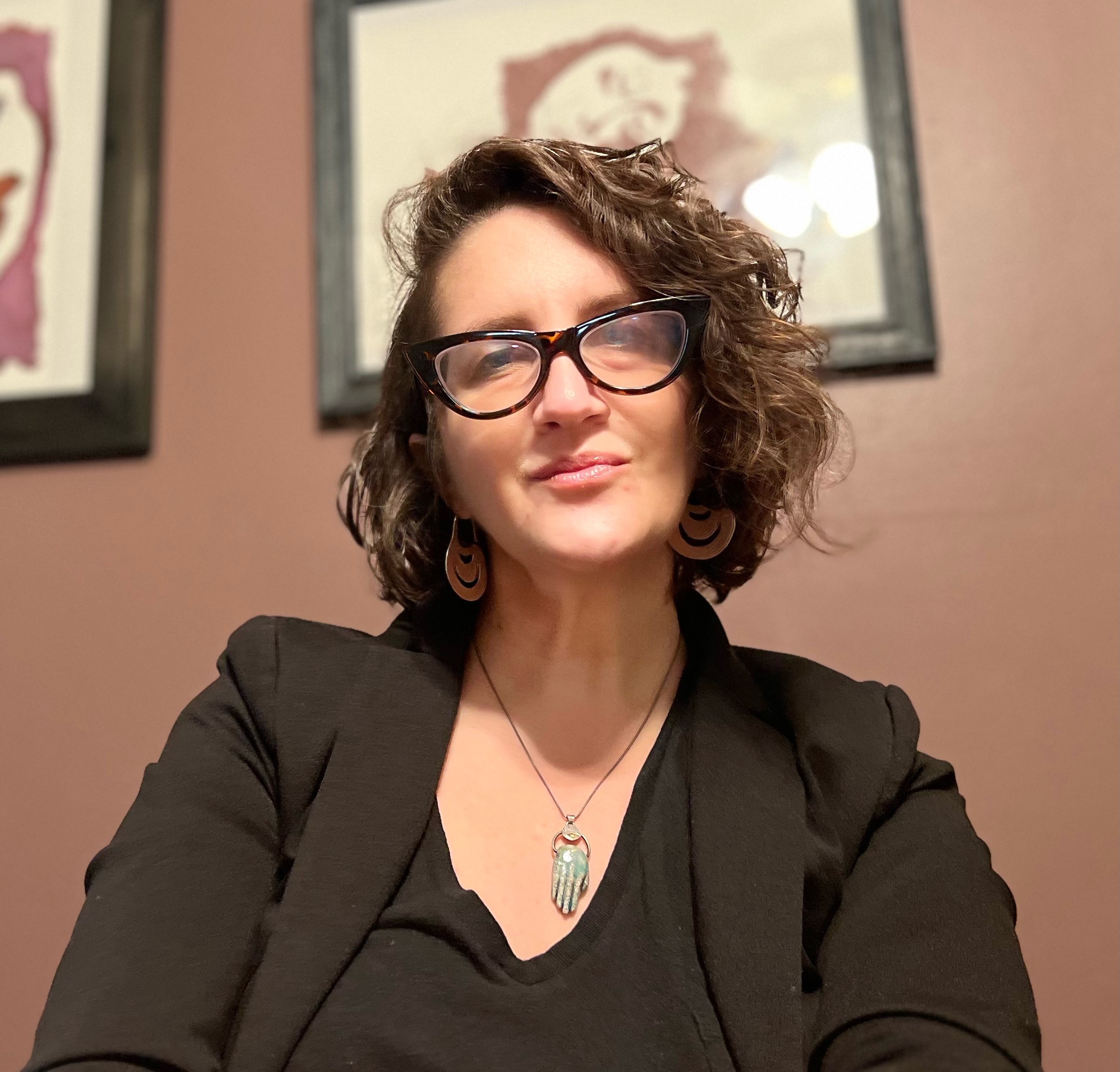 Image of Dr. Alaina Brenick in a black top and black blazer with tortoise shell glasses in a cat eye shape. She is wearing a light blue batique Hamsa necklace. She has short brown wavy hair, slightly shaved on the right side. 