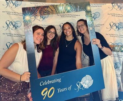 Picture of four scholars in front of a white backdrop with "SPSSI The Society for the Psychological Study of Social Issues" written in blue. The scholars hold up a frame around their faces that says "Celebrating 90 Years of SPSSI"